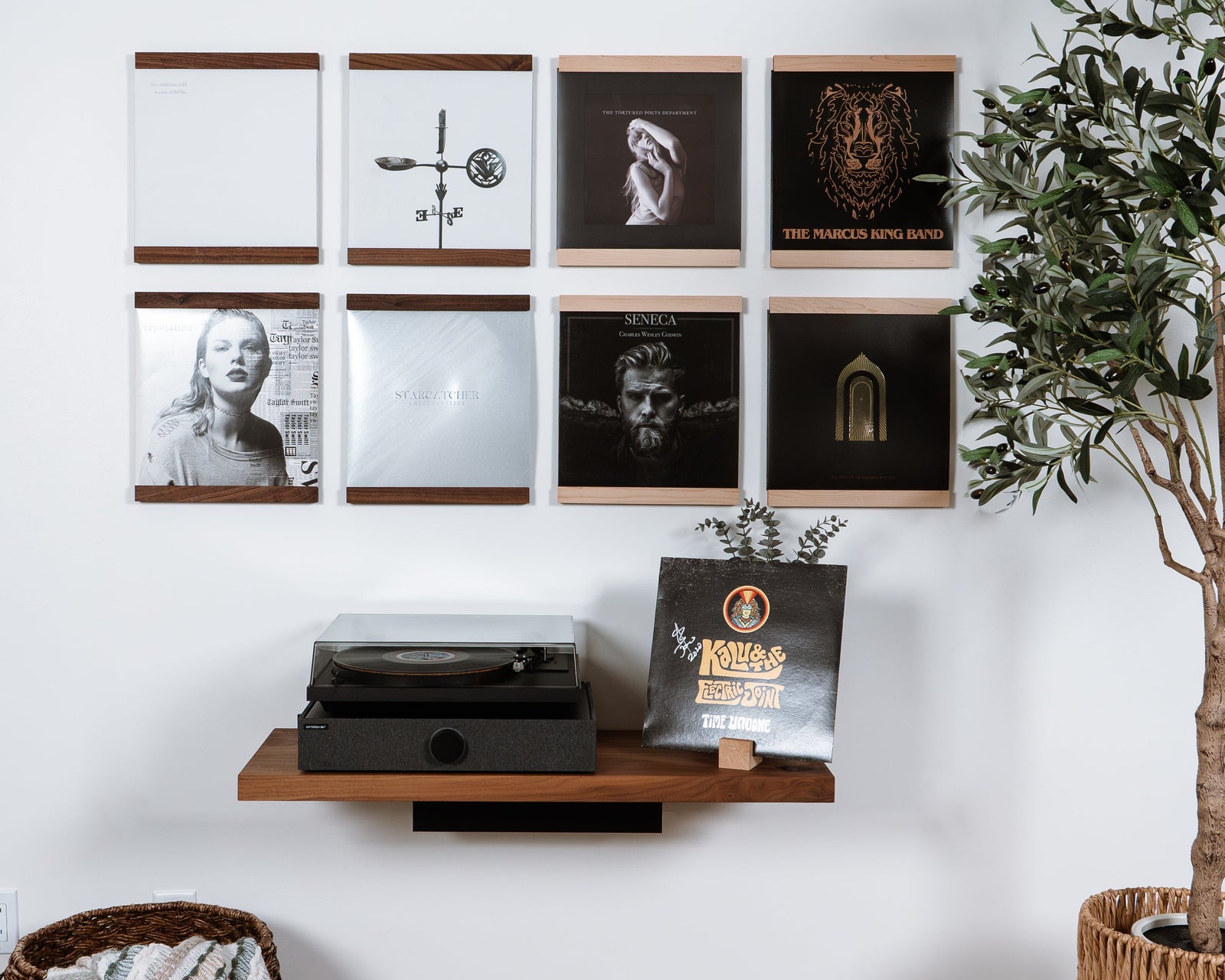 A styled interior wall showing a wall-mounted floating wooden shelf in Walnut and Maple hardwoods that trim the top and bottom of a vinyl record. Eight units organized in a grid above a record player. The product is the wall shelf itself, shown in a real room with wood flooring, a plant, a basket with a throw blanket. 