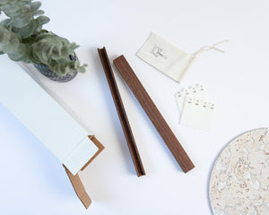 A styled flat-lay of a product set on a white background, featuring the two walnut pieces that come with every unit which are the top and bottom of the Drop-In Display, a small cream cotton pouch with the Deep Cut logo stamped on it and eight velcro adhesive stickers for mounting. A cork slip mat, a vase of eucalyptus and a white box are partially visible as props. 